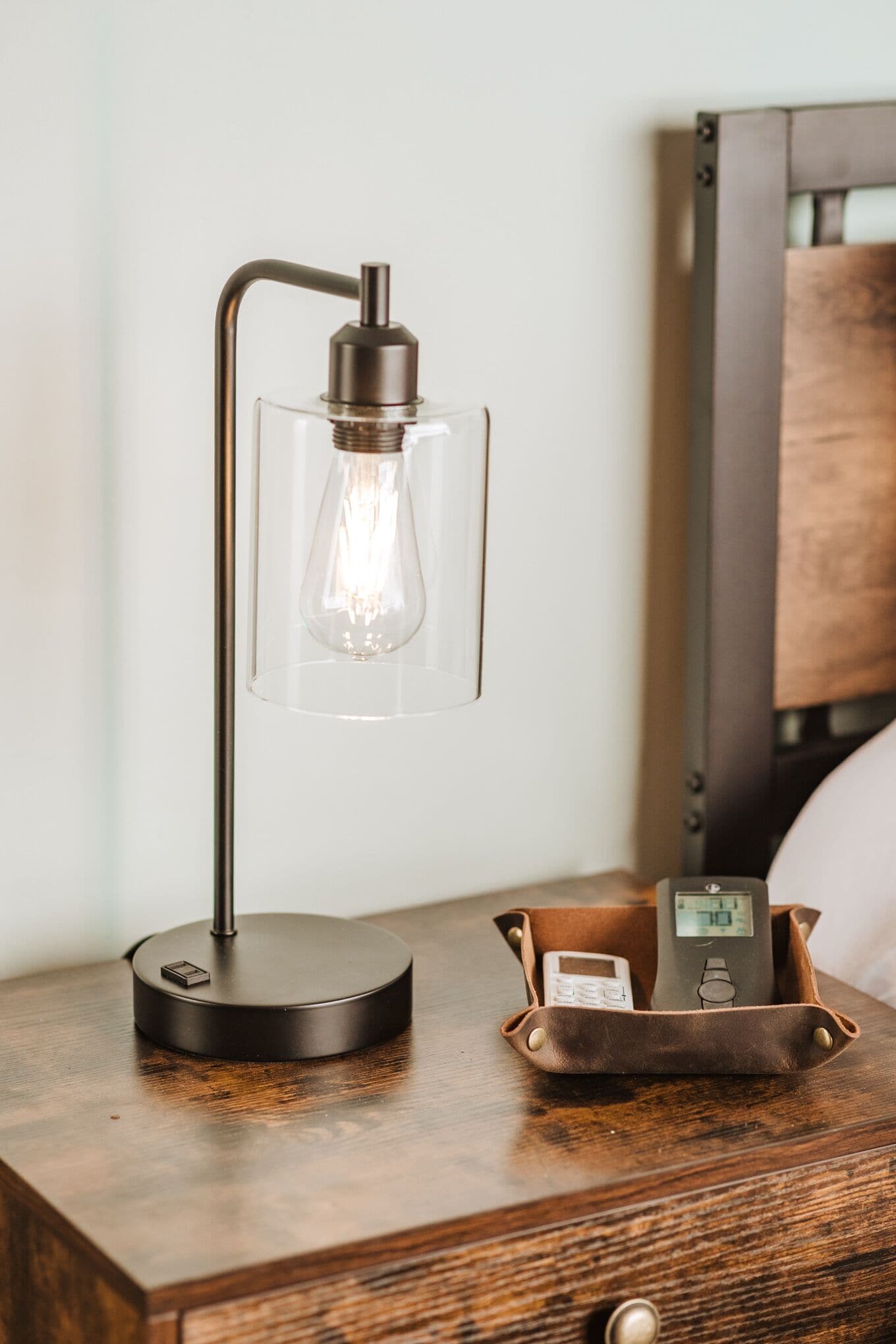 Ett sängbord med en modern lampa, en digital klocka och en läderpåse.