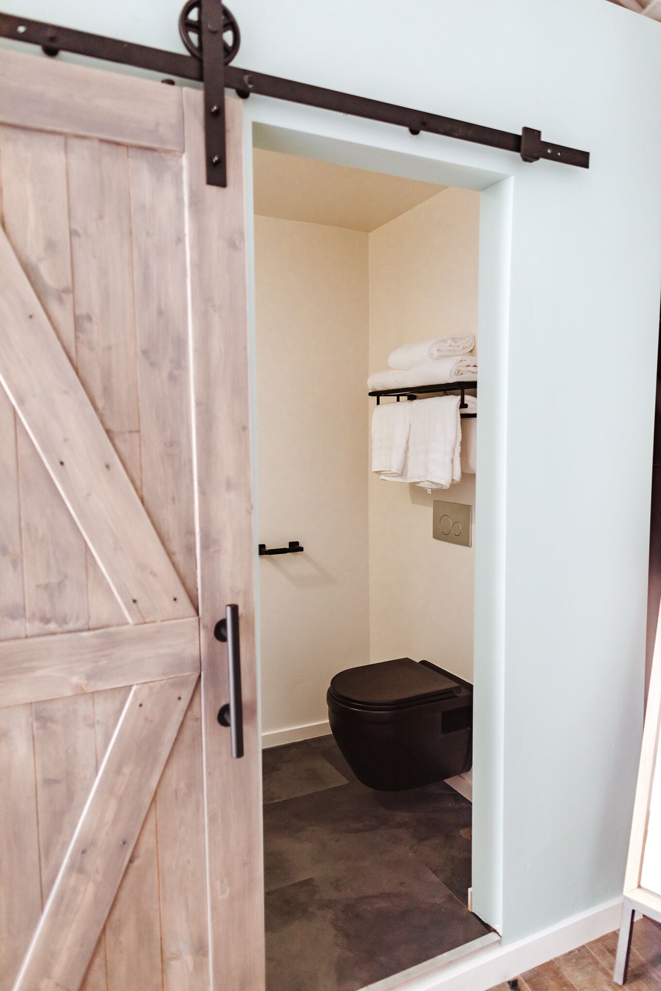 Un baño con una puerta de madera, un cesto de basura negro y un perchero con toallas dobladas.