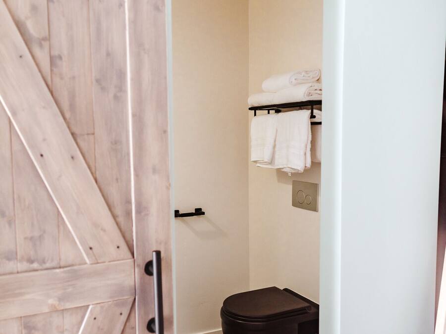 Un baño con una puerta de madera, un cesto de basura negro y un perchero con toallas dobladas.