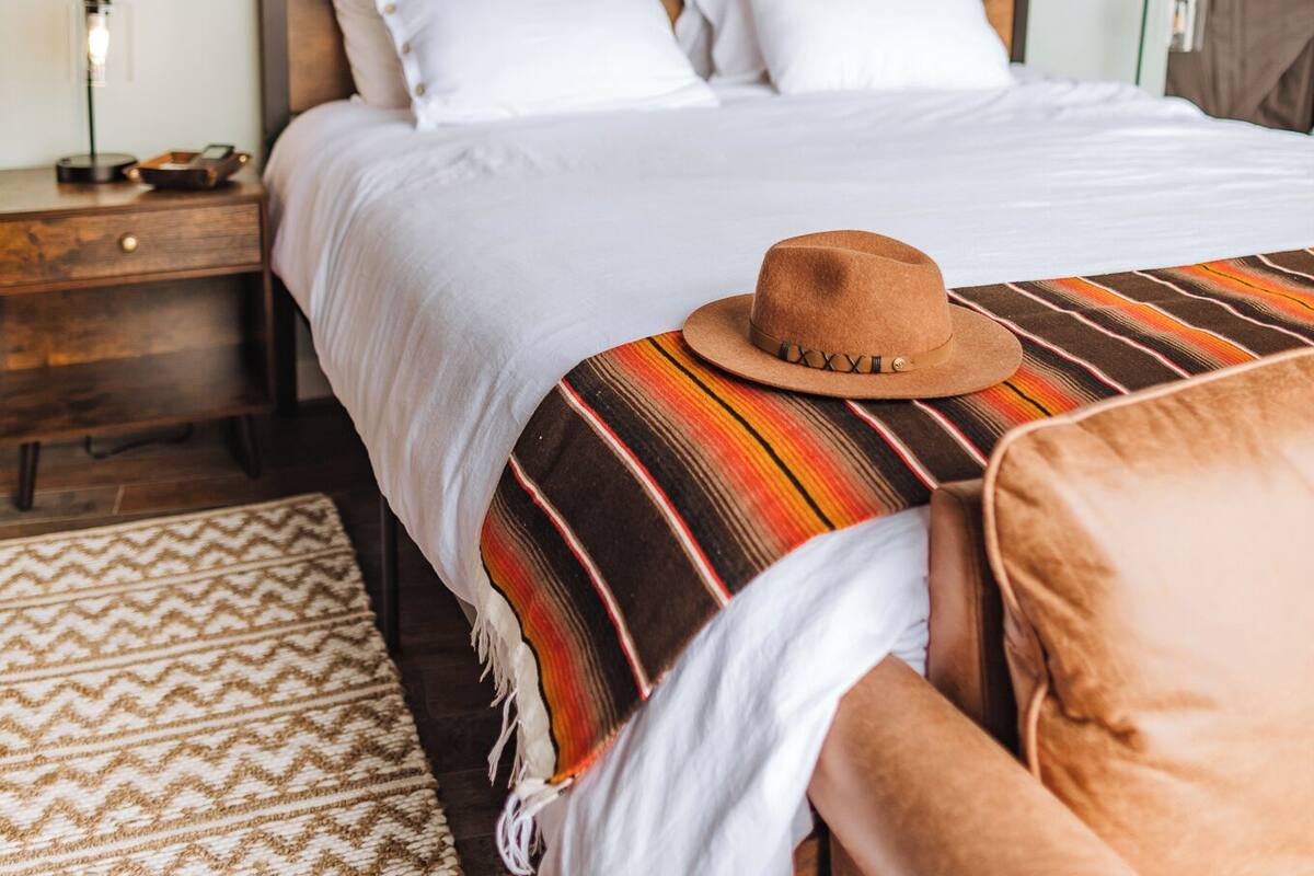 A neatly made bed with a brown hat on it, a bedside table with a lamp, and a wall hanging above the bed.