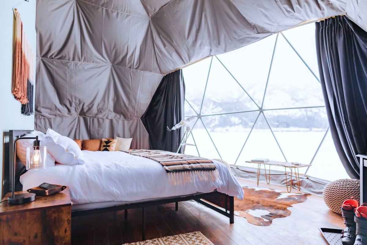 A cozy bedroom with a large window, a wooden bedside table, and a bed with a patterned blanket.