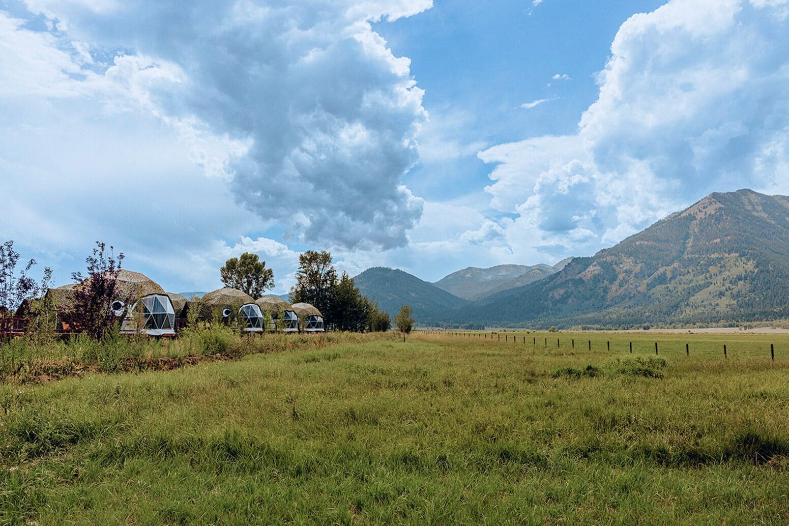 Paisaje rural con una hilera de estructuras de cúpulas geodésicas, un campo cercado y montañas al fondo.