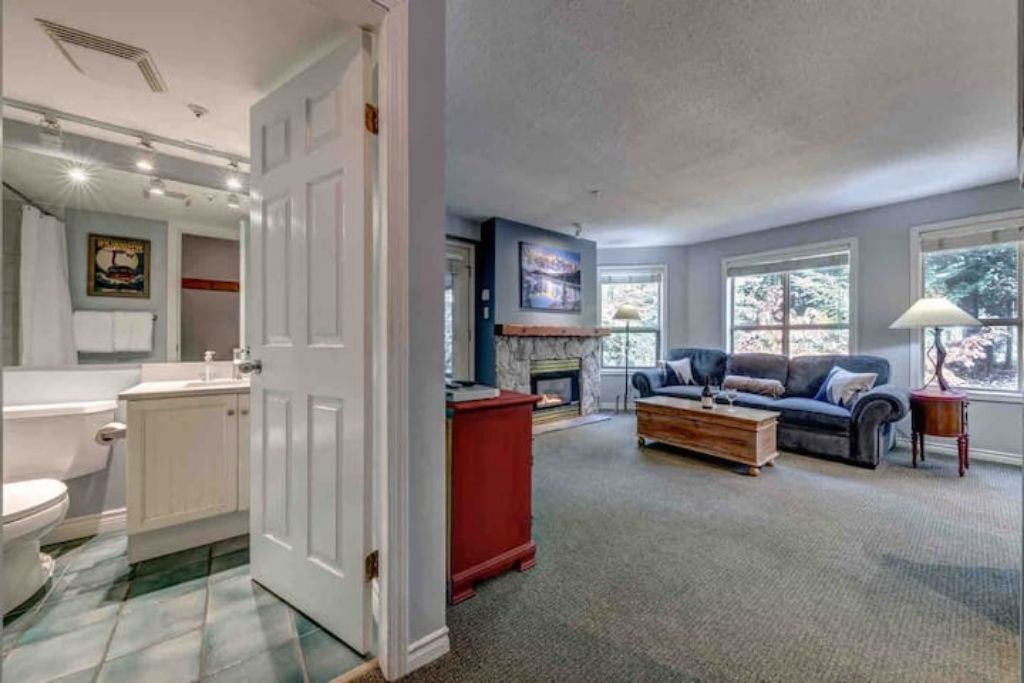 A living room with a fireplace, a sofa, and a coffee table. There is a door leading to a bathroom with a toilet and sink.