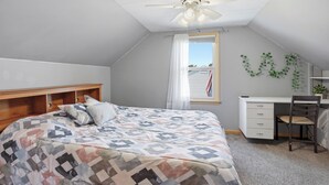 Quarto com cama, escrivaninha, cadeira, ventilador de teto e janela com cortinas.