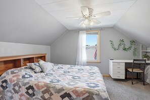 Un dormitorio con cama, escritorio, silla, ventilador de techo y ventana con cortinas.