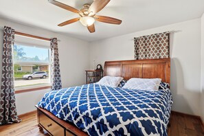 A bedroom with a wooden bed, a ceiling fan, a window with curtains, and a view of the street.