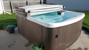 Uma banheira de hidromassagem com revestimento de madeira e tampa branca, em superfície de concreto com grama e plantas em vasos por perto.