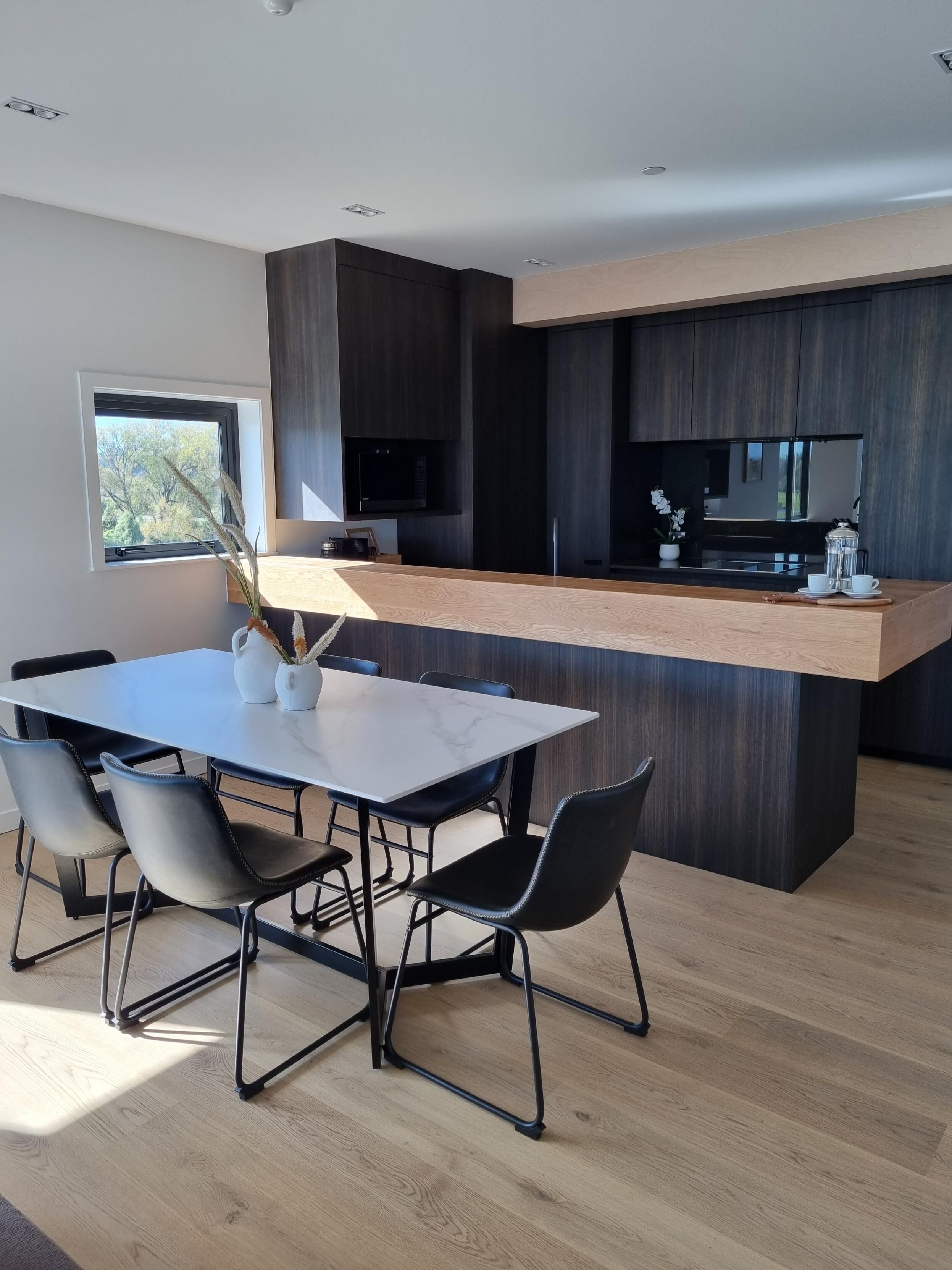 Une cuisine moderne dotée d’un îlot central, de meubles de rangement aux couleurs sombres et d’un coin repas avec une table en marbre et des chaises noires.