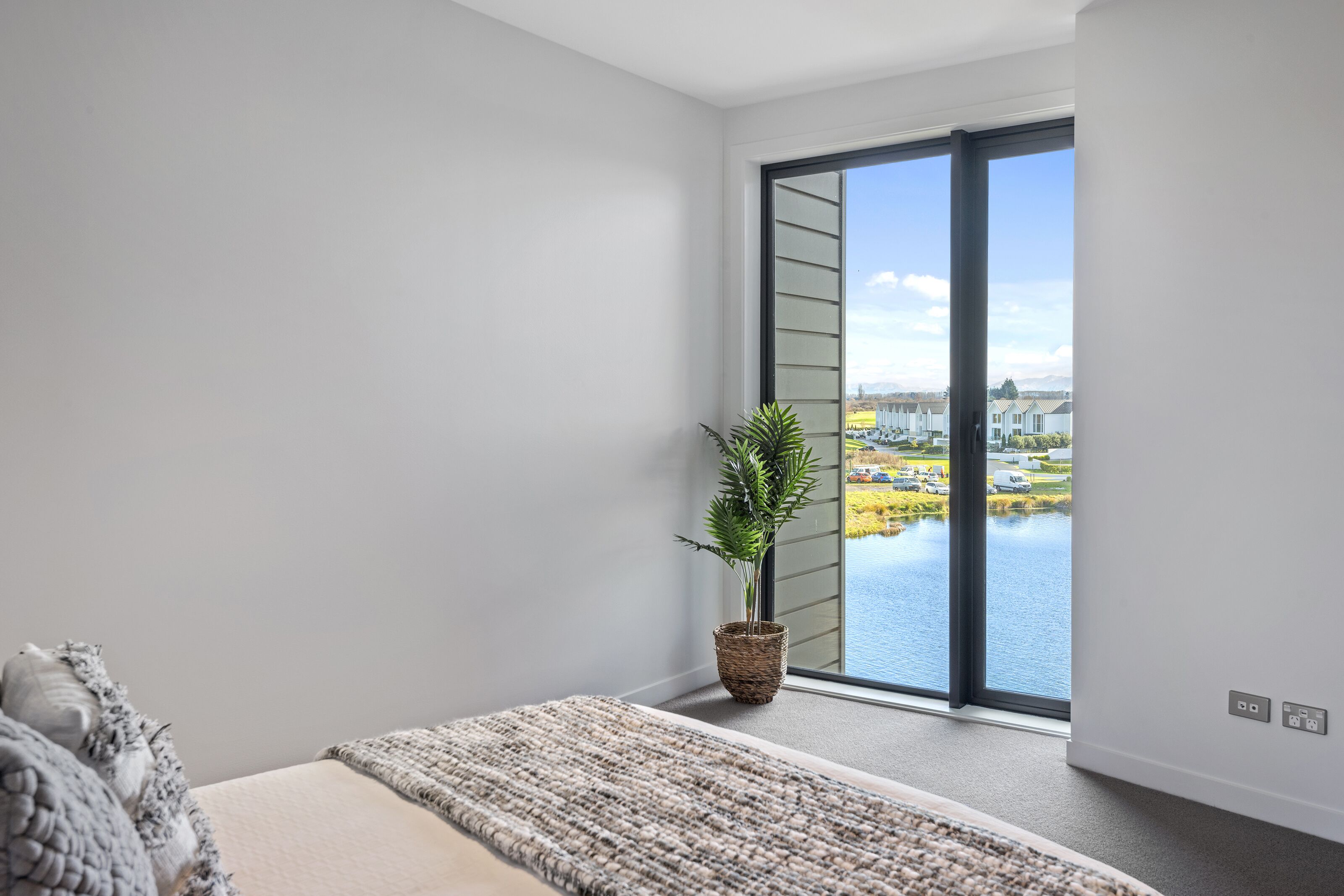 A modern bedroom with a large window, a bed, a plant, and a view of a body of water and buildings.