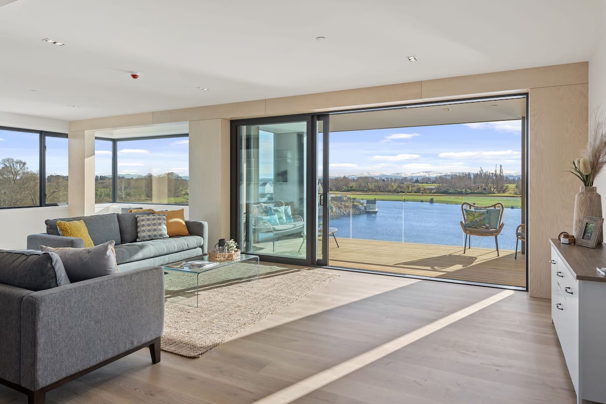 A spacious living room with a large sliding glass door leading to a balcony overlooking a lake and mountains.
