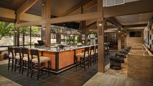 A spacious bar area with high ceilings and large windows, featuring a bar counter with stools and a seating area with round tables.