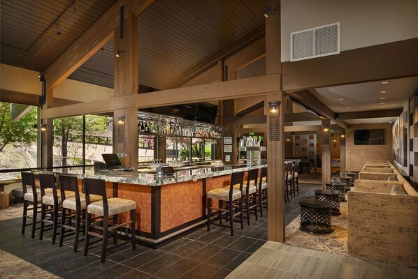A spacious bar area with high ceilings and large windows, featuring a bar counter with stools and a seating area with round tables.