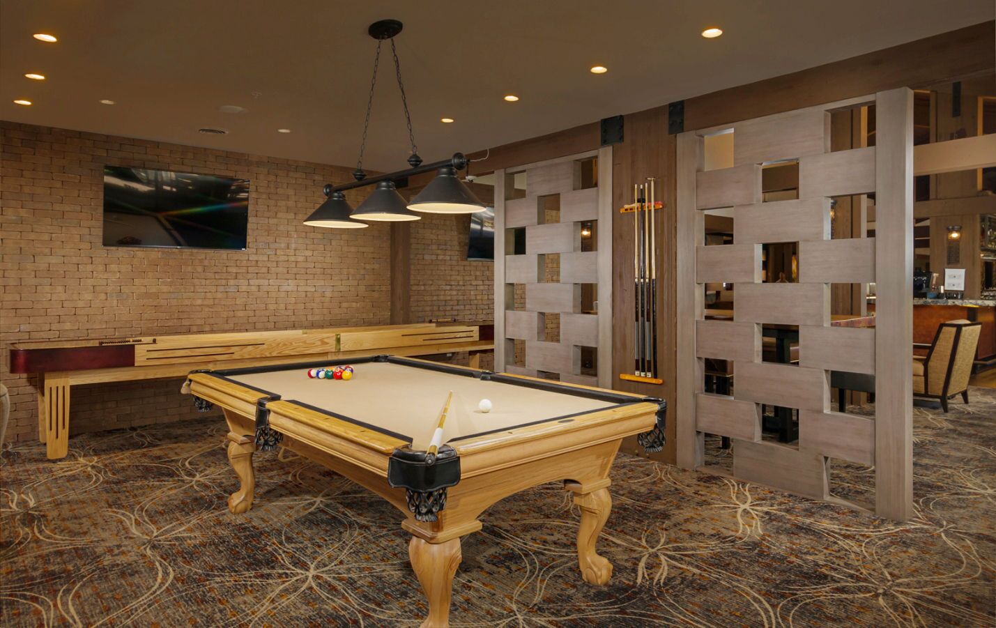 A pool table with a billiard cue, a pool table, and a billiard rack in a room with a patterned carpet and brick wall.