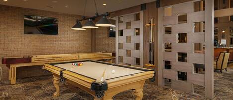 A pool table with a billiard cue, a pool table, and a billiard rack in a room with a patterned carpet and brick wall.