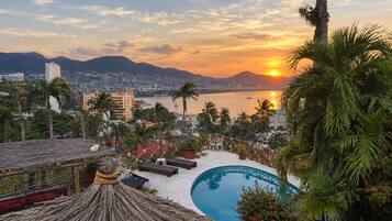 Un resort con piscina, sillas de descanso y vistas al atardecer sobre una ciudad y montañas.