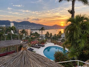 A resort with a pool, lounge chairs, and a view of the sunset over a city and mountains. - Casa Breizh (Acapulco)