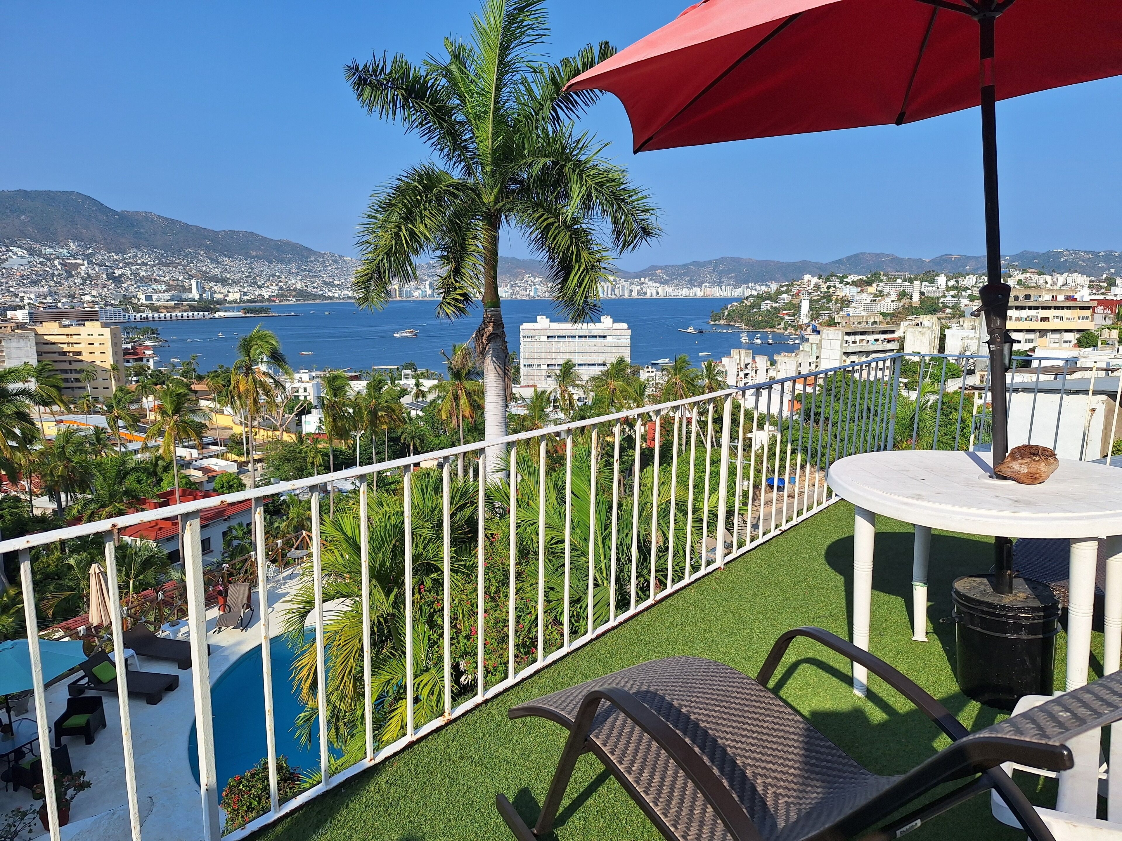 A balcony with a view of the sea, a pool, and a red umbrella.