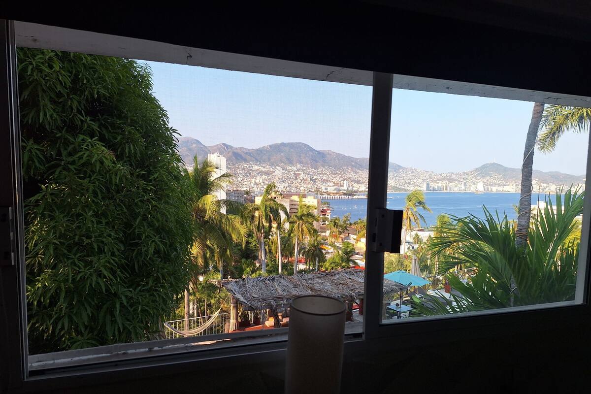 A room with a large window overlooking a coastal cityscape, including a beach and mountains.