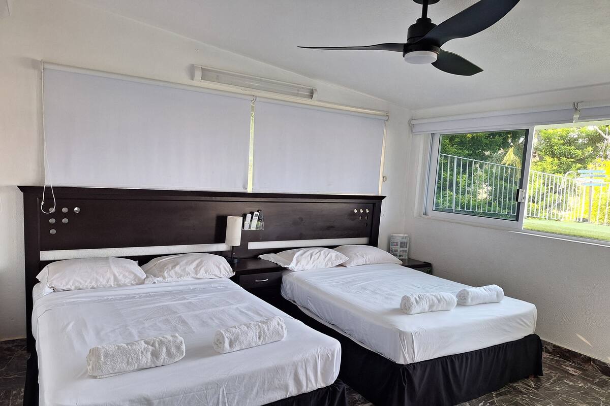 A hotel room with two beds, a ceiling fan, a window with a view of greenery, and a bedside table with a lamp.