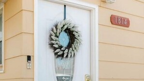 A white door with a wreath, a doormat with a message, and a house number sign.