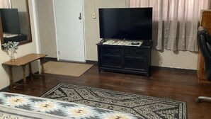 Ein Wohnzimmer mit einem Flachbildfernseher auf einer schwarzen Kommode, einer hölzernen Bank, einem gemusterten Teppich, einer weißen Tür und einem Vorhang.