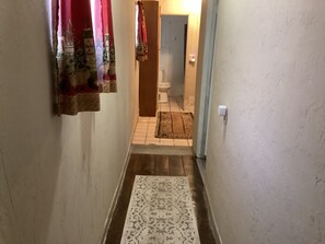 Un couloir étroit, orné d’un tapis à motifs, avec une porte en bois et des toilettes visibles au fond.