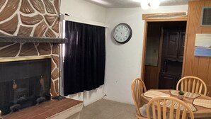 Ein Essbereich mit rundem Tisch und Stühlen, einem Kamin mit Steinmuster, einer Wanduhr und einer Tür zu einem anderen Raum.
