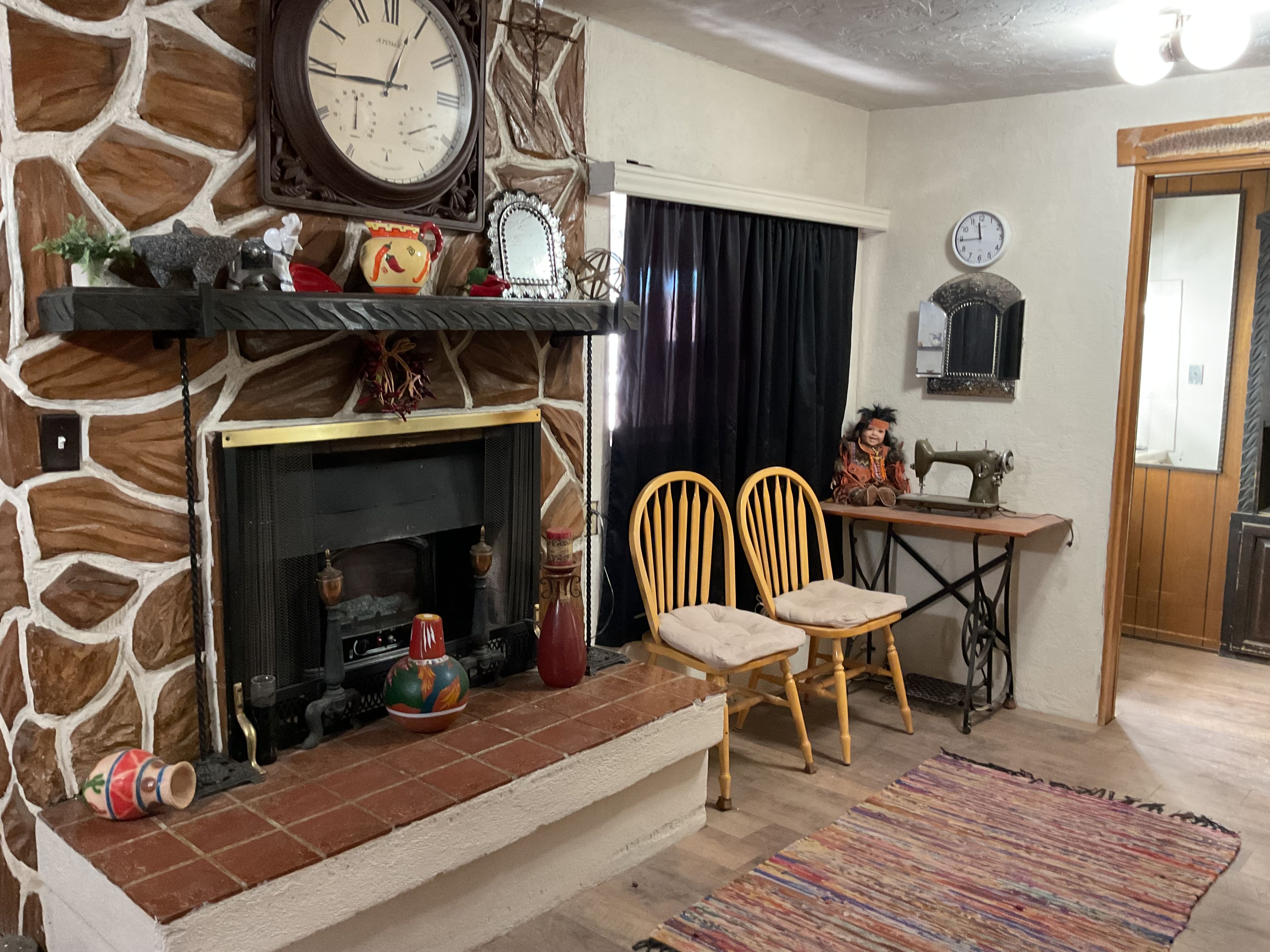 A room with a stone wall, a fireplace, two wooden chairs, a sewing machine, and a clock on the wall.