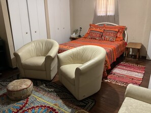 Une chambre à coucher comprenant un lit, deux fauteuils, une table de chevet, une commode, une lampe, une fenêtre avec des rideaux et un tapis à motifs.