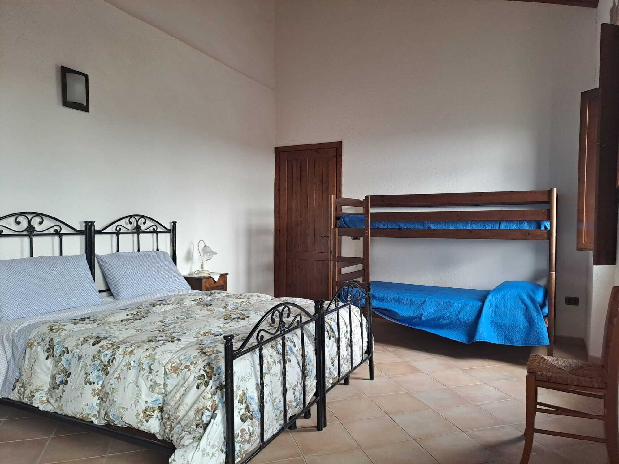 A bedroom with a single bed, a bunk bed, a wooden wardrobe, a chair, and a small table with a lamp.