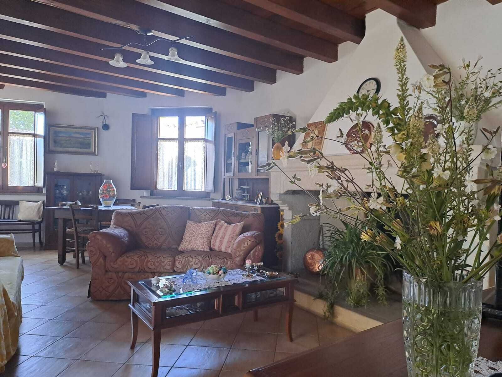 Un salón acogedor con un sofá, una mesa de centro y un jarrón grande con flores. Hay una ventana con cortinas, una chimenea y una zona de comedor al fondo.