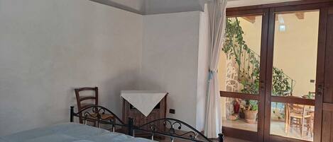 A bedroom with a bed, a chair, a small table, and a sliding glass door leading to an outdoor area with plants.