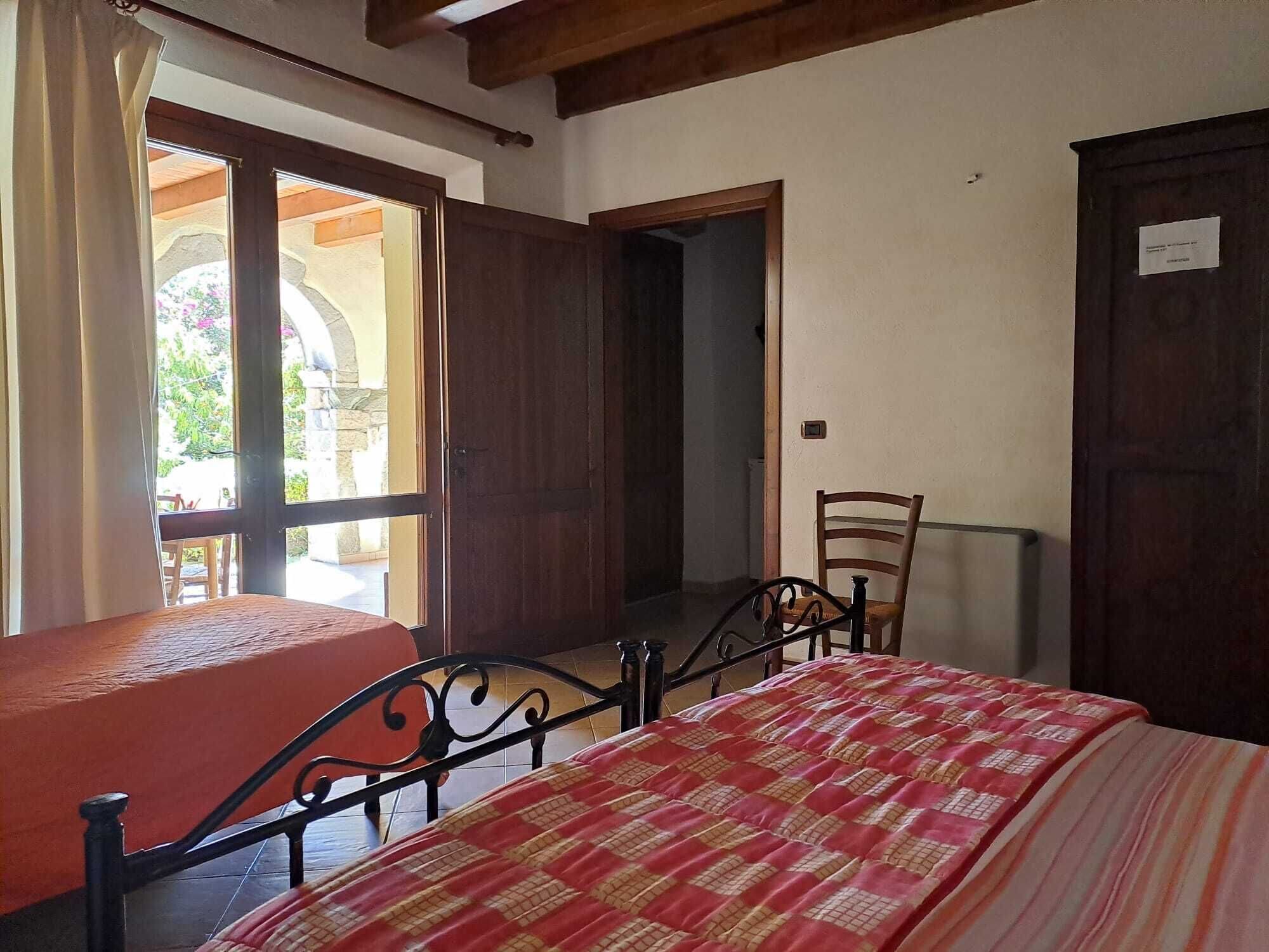 Habitación con cama, una silla y una mesa. Tiene una ventana con cortinas, una puerta y vistas al exterior.