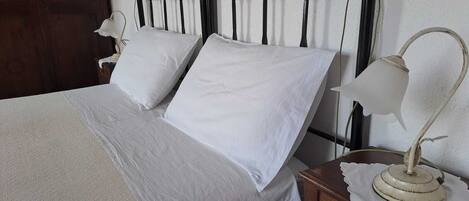 Una cama con sábanas blancas, una mesita de noche de madera con una lámpara y una pared de diseño sencillo.