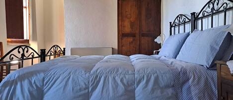A bed with a white duvet and blue patterned pillows, a wooden wardrobe, and a stone wall.