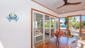 Zona pranzo con tavolo e sedie in legno, ventilatore a soffitto e vista sulla spiaggia attraverso ampie porte in vetro.