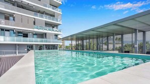 A swimming pool with clear water, surrounded by a modern building with balconies and a covered area.
