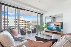A modern living room with a large TV, comfortable seating, a balcony with a city view, and a potted plant. - Resort-style Getaway for Groups by the Marina (Cairns City)