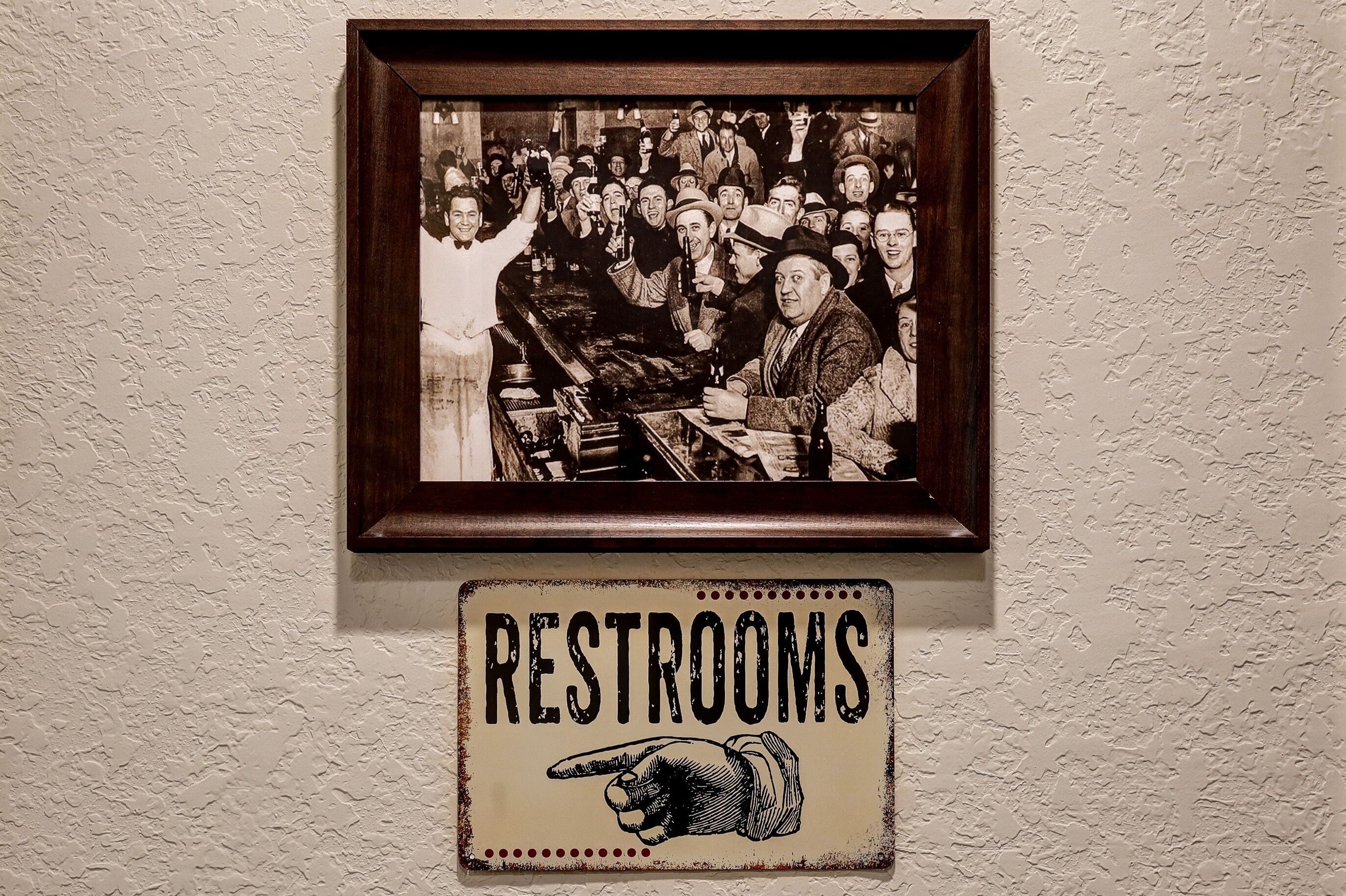 A framed photograph of a crowded bar scene with a sign below reading "RESTROOMS" and a pointing hand graphic.
