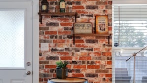 A room with a brick wall, a wooden barrel, a potted plant, and a shelf with bottles and a sign. - Speakeasy - A Birdy Vacation Rental (San Antonio)