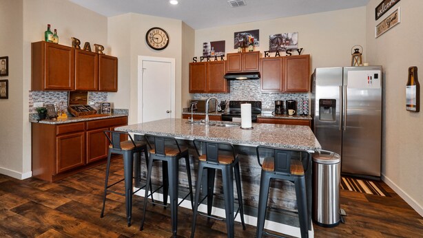 A kitchen with wooden cabinets, a stainless steel refrigerator, a granite countertop, and a bar area with stools. - Speakeasy - A Birdy Vacation Rental (San Antonio)