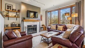 A living room with leather sofas, a fireplace, a TV, and large windows with a view of trees. - Beautifully upscale condo with pool, hot tub, and fitness room Highridge E1 (Killington)