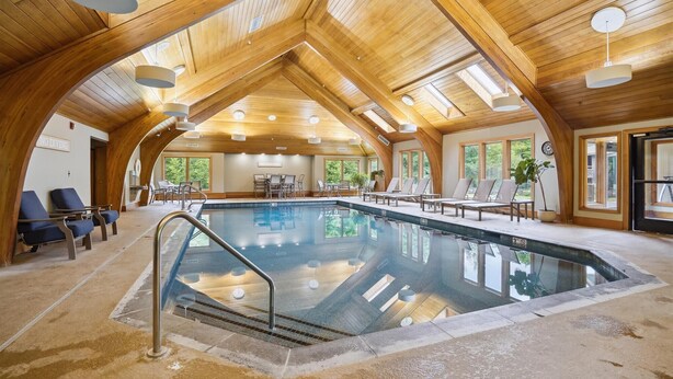 An indoor swimming pool with a wooden ceiling, lounge chairs, and a view of the outdoors. - Beautifully upscale condo with pool, hot tub, and fitness room Highridge E1 (Killington)