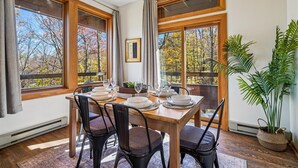 A dining area with a wooden table set for four, large windows with curtains, and a potted plant on the right. - Beautifully upscale condo with pool, hot tub, and fitness room Highridge E1 (Killington)