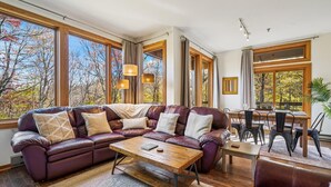 A spacious living room with a leather sectional sofa, a wooden coffee table, and a dining area with a wooden table and chairs. Large windows provide views of trees and a clear sky. - Beautifully upscale condo with pool, hot tub, and fitness room Highridge E1 (Killington)