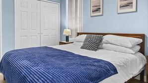 Un dormitorio con una cama, almohadas blancas, una colcha azul, un cabecero de madera, una mesita de noche con una lámpara, un ventanal con persianas y dos cuadros enmarcados en la pared.