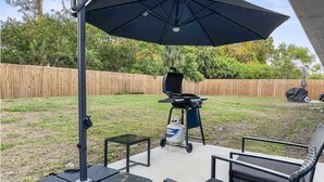 Un patio con una parrilla, mobiliario exterior y un paraguas grande.