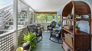 A covered porch with wicker rocking chairs, a table, and a wooden shelf with baskets and a potted plant. - Sandcrab - Quaint Ocean View Cottage, Beach Access Directly Across (Folly Beach)