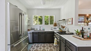 A modern kitchen with dark cabinets, a stainless steel refrigerator, a microwave, and a stove. There are windows providing natural light and a view of greenery outside. - Sandcrab - Quaint Ocean View Cottage, Beach Access Directly Across (Folly Beach)