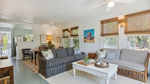 A spacious living room with a white ceiling fan, striped sofas, a white coffee table, and large windows with bamboo blinds. - Sandcrab - Quaint Ocean View Cottage, Beach Access Directly Across (Folly Beach)